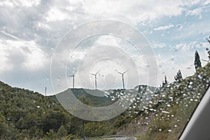 Wind turbines under a cloudy sky seen through the wet glass window of the car