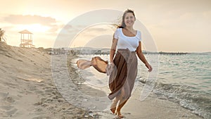 Wind whips woman's scarf as she joyfully dances and runs on the sandy beach at sunset. Highlights the concepts of fun,