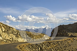 A winding asphalt road in the desert mountains against the snowy peaks under a blue sky with white clouds