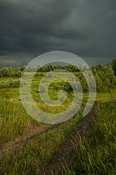 A winding, dirt rural road running through a meadow lit by sunlight, and above it a stormy sky with leaden storm clouds
