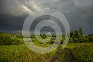 A winding, dirt rural road running through a meadow lit by sunlight, and above it a stormy sky with leaden storm clouds