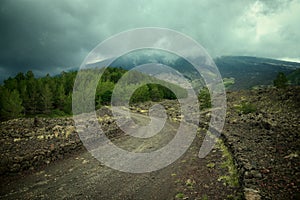 Windirg dirt road along the slopes of Etna Mount leading to the tree line under stormy sky