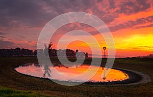 Windmill by pond in rural field with vivid sunrise