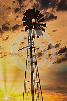 A windmill silhouette at sunset with the sun and moon and cloudy sky