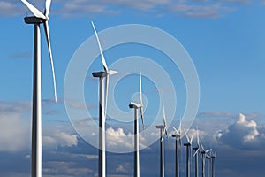 Windmills generating electricity stand in a row against a blue sky with clouds on a beautiful bright day.