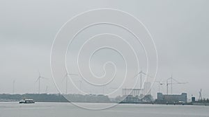 Windmills with white blades against river and industrial city at foggy morning