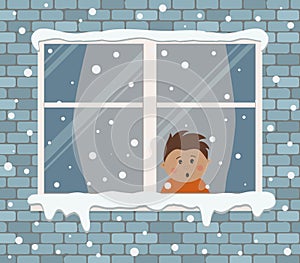 Window on a brick wall on a snowy day. A little boy in the room is surprised, looking at the snow