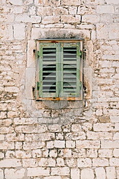 A window with closed, old, green, wooden shutters in the brick wall of the house.