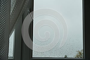 A window with curtains covered with rain drops of a heavy downpour. Cloudy dark grey sky is on the background.