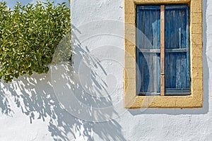 The window of a Greek house on a white wall with yellow trim and blue wooden shutters