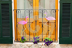 Window with green shutters, decorated with two pink flamingos and some flower pots with pink petunia