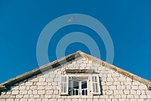 A window with open shutters under the roof of a house against a bright blue sky with a seagull in flight.