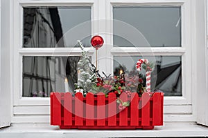 Window sill with Christmas decoration in red flower box. Small Christmas tree, red ball and red and white candy cane.