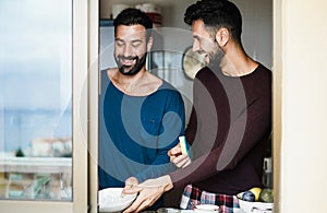 Window view of gay couple washing dishes together inside home kitchen - Young hosexual people having fun during morning routine -