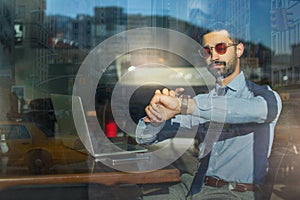 Through window view of handsome businessman wearing businesswear and eyeglasses checking time on his wrist watch at cafe