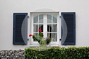 A window with white frames, dark blue shutters and flowers on the windowsill. A facade of a traditional European country house