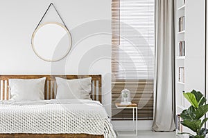 Window with wooden blinds and light grey curtain in white bedroom interior with mockup poster, double bed and rack with books