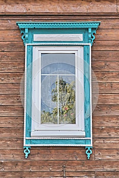 Window with wooden carved turquoise white architraves on brown plank facade of old house.