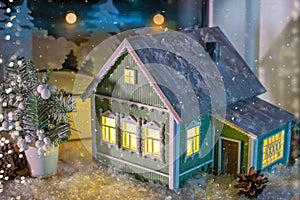 On windowsill, among Christmas tree decorations, night light in form of village house glows, against background of night snow