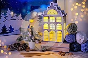 On windowsill, among the skeins of yarn, night light in form of an old European house glows against background of night snow