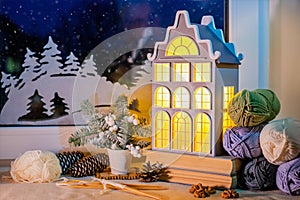 On windowsill, among the skeins of yarn, night light in form of an old European house glows against background of night snow