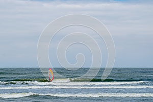 Windsurfer in black suit controls board with red and yellow sail from foaming waves rushing ashore deep in sea to strip of horizon