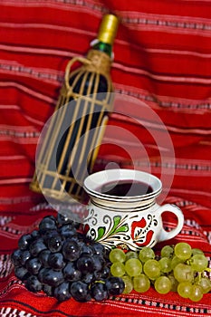 Wine bottle, traditional mug filled with red wine and red and white grapes on a traditional Romanian carpet