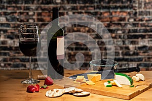 Wine and a glass accompanied by cheese on a table with a cutting board and cutlery with fruit.