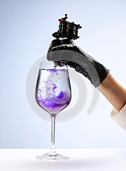 Wine glass and purple ink. Tattooer master& x27;s hand in black glove holding machine for making tattoo art on body