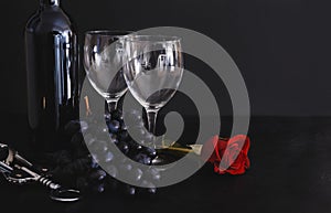 2 wine glasses, a bottle of red wine, a bunch of dark grapes, a metal corkscrew, a red rose on a black background