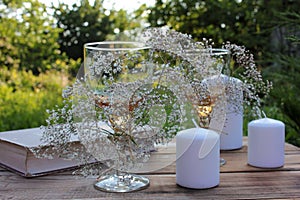 Wine glasses decorated with flowers, old book, and candles on a wooden table on a blurred green background in summer garden