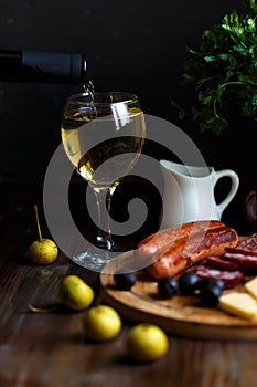 Wine pours into a glass goblet. On the table, meat appetizer, fried sausages, salami, cheese, olives.