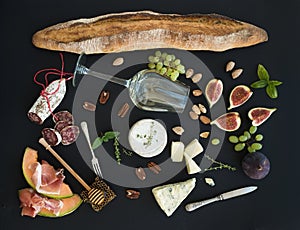 Wine and snack set. Baguette, glass of white, figs, grapes, nuts, cheese variety, meat appetizers, herbs on black grunge