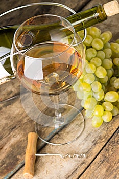 Wineglass with white wine, botlle, corkscrew and cluster of grapes around on wood background