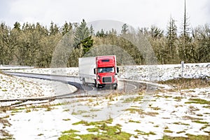 Winer landscaping with red big rig semi truck with dry van semi trailer turning on the exit from highway road between hills