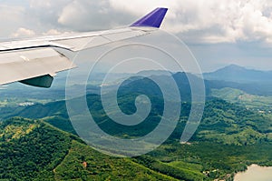 Wing of airplane flying in cloudy sky over the green mountains.