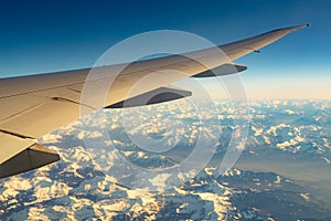 Wing of plane over mountain cover with white snow. Airplane flying on blue sky. Scenic view from airplane window. Commercial