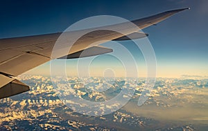 Wing of plane over mountain cover with white snow. Airplane flying on blue sky. Scenic view from airplane window. Commercial