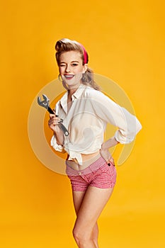 Winking. Beautiful young blonde girl in white shirt and pink shorts posing with tool, wrench against yellow studio