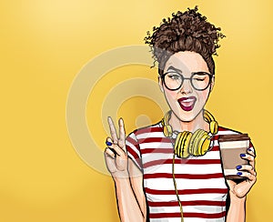 Winking woman in glasses with head phones makes peace gesture  Pop art girl holding coffee cup.