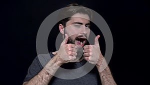 Winking young man with beard showing thumb up. Cheerful man approvingly winks and shows his thumb up. Human facial