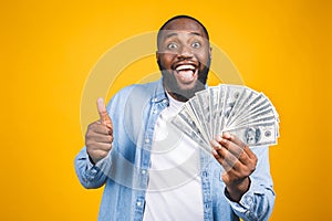 Winner! Young rich african american man in casual t-shirt holding money dollar bills with surprise isolated over yellow wall.