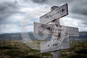 Winners never quit text on wooden rustic signpost outdoors in nature/mountain scenery.