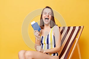 Winning journey. Long awaited travel. Overjoyed attractive woman wearing striped swimming suit sitting on deck chair with passport