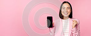 Winning korean businesswoman showing smartphone screen, smiling pleased, demonstrating mobile phone application, online