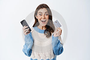Winning money online. Excited girl holding plastic credit card and mobile phone and smiling amazed, staring at camera