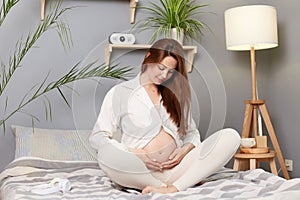 Winsome adorable pregnant woman touching her bare belly wearing white clothing sitting in bed, enjoying pregnancy, resting in