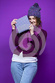 Winsome Adult Caucasian Woman in Warm Knitted Hat and Purple Scarf Posing with Big Wrapped Giftbox Whie Smiling Happily With Eyes