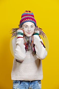 Winsome Caucasian Blond Girl In Warm Knitted Hat and Scarf Posing with Lifted Hands While Touching Cheeks With Knitted Color Scarf