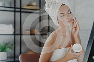 Winsome pleased young European woman puts cream on face, looks at her reflection in mirror, wrapped in bath towel, has anti aging